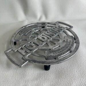 May include: A silver-colored metal trivet with the words "HOT DINING" in large letters. The trivet has a circular design with a small black foot. Text on the trivet reads "Warmth in the your kettle makes your cooking excellent. 1952 KITCHEN EQUIPMENT AND APPLIANCES."