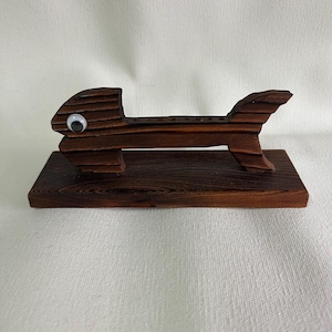 Puede incluir: Un objeto artesanal de madera con forma de pez y tinte marrón oscuro. El pez tiene una base rectangular y un cuerpo con una serie de pequeños agujeros. Un ojo saltones está unido al lado de la cabeza del pez. La veta de la madera es visible.