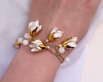 May include: A gold bracelet with white flower charms and white pearl beads. The bracelet is on a person's wrist.