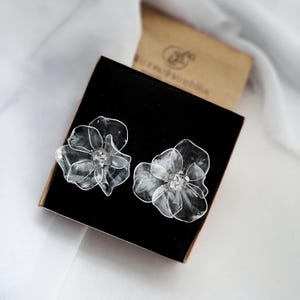 May include: Two clear, flower-shaped earrings with a central crystal detail, displayed in a black box. The earrings feature delicate, translucent petals, set against a white background.