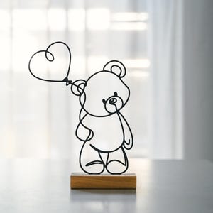 Könnte beinhalten: Eine schwarze Drahtskulptur eines Teddybären, der einen herzförmigen Ballon hält. Die Skulptur steht auf einem Holzsockel.