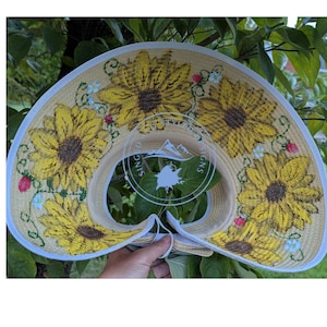May include: A straw visor with a floral sunflower design. The brim is decorated with embroidered yellow sunflowers, small blue flowers, and red berries. The visor has a white trim and a clear plastic insert.