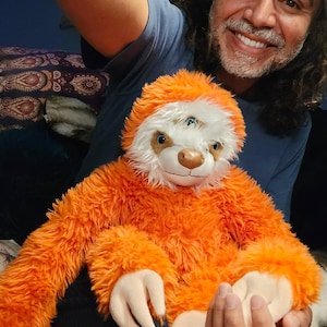 Puede incluir: Una persona sostiene un gran perezoso de peluche naranja y blanco con una cara sonriente. El perezoso tiene tres ojos y un pelaje largo y esponjoso.