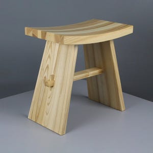 Peut inclure: Un tabouret en bois clair avec une assise incurvée et des pieds inclinés. Le tabouret présente un joint en forme de croix sur un côté et une barre de support horizontale. Le grain du bois est visible.