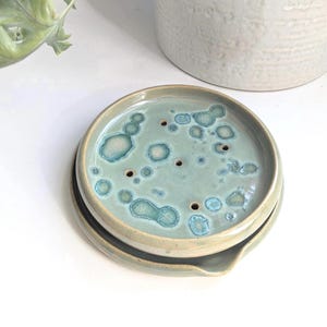 Könnte beinhalten: Eine Keramik-Seifenschale mit hellgrüner Glasur und einem gesprenkelten Muster. Die Schale hat eine runde Form und einen kleinen Rand.