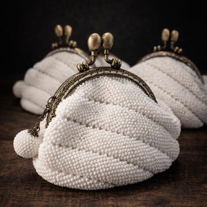 May include: Three white beaded coin purses with antique bronze clasps and frames on a dark wooden surface. The purses have a textured, wave-like design and a small beaded ball attached to a chain.