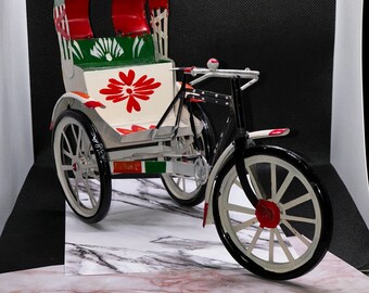 May include: A decorative miniature rickshaw model. The rickshaw features a red and white canopy, a white body with a red floral design, and black wheels with white rims. The model is made of metal and has a detailed design.
