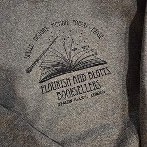 May include: Gray sweatshirt with a black graphic featuring an open book, wand, and text. The text reads "SPELLS HISTORY FICTION POETRY PROSE" and "FLOURISH AND BLOTTS BOOKSELLERS DIAGON ALLEY, LONDON". The establishment date "EST. 1654" is also included.
