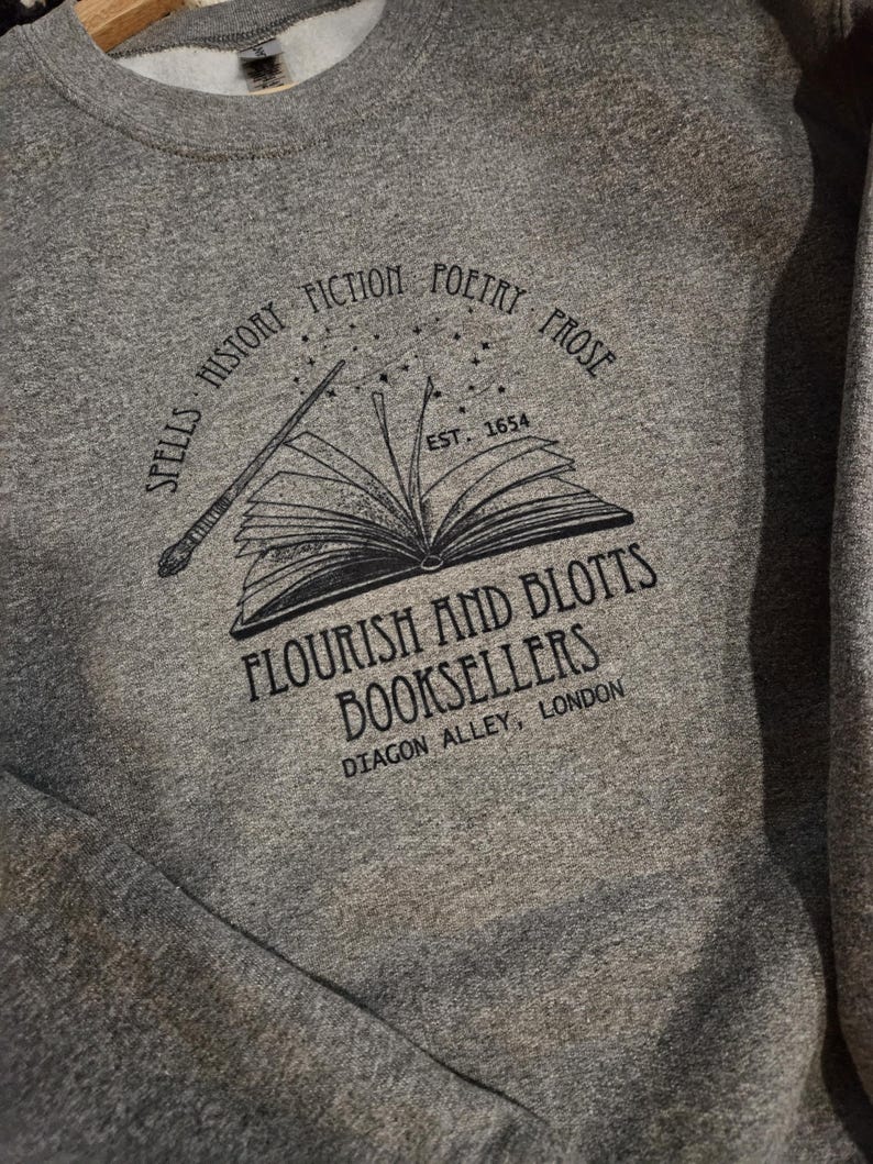 May include: Gray sweatshirt with a black graphic featuring an open book, wand, and text. The text reads "SPELLS HISTORY FICTION POETRY PROSE" and "FLOURISH AND BLOTTS BOOKSELLERS DIAGON ALLEY, LONDON". The establishment date "EST. 1654" is also included.