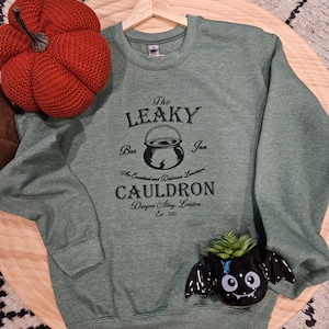 May include: Green sweatshirt with a black graphic of The Leaky Cauldron, a bar and inn in Diagon Alley, London. The graphic includes a cauldron with a lightning bolt and the text "An Excellent and Delicious Luncheon".