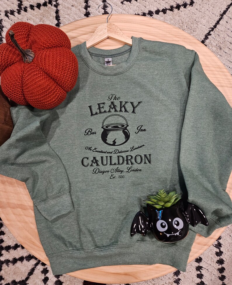 May include: Green sweatshirt with a black graphic of The Leaky Cauldron, a bar and inn in Diagon Alley, London. The graphic includes a cauldron with a lightning bolt and the text "An Excellent and Delicious Luncheon".