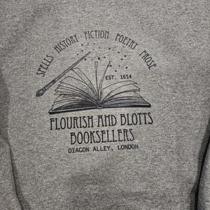 May include: A heather gray sweatshirt featuring a black graphic of an open book with a wand and stars. The text around the book reads "Spells, History, Fiction, Poetry, Prose" and "Flourish and Blotts Booksellers, Diagon Alley, London".