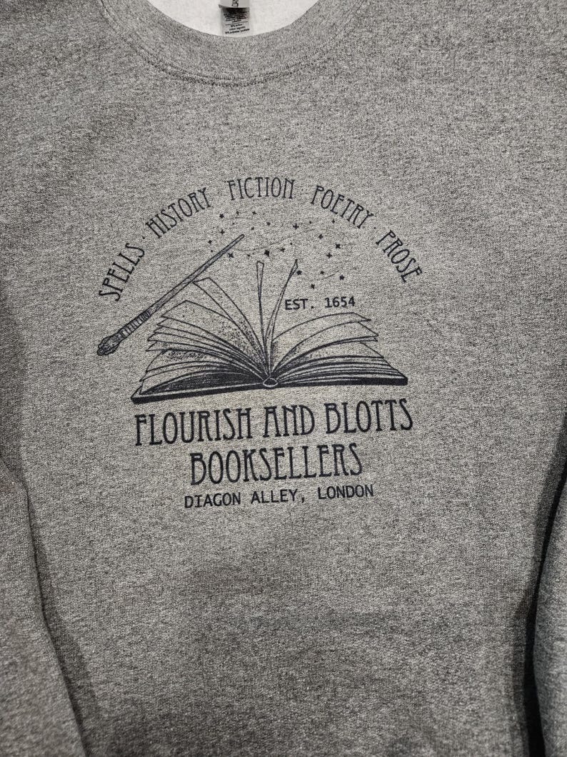 May include: A heather gray sweatshirt featuring a black graphic of an open book with a wand and stars. The text around the book reads "Spells, History, Fiction, Poetry, Prose" and "Flourish and Blotts Booksellers, Diagon Alley, London".