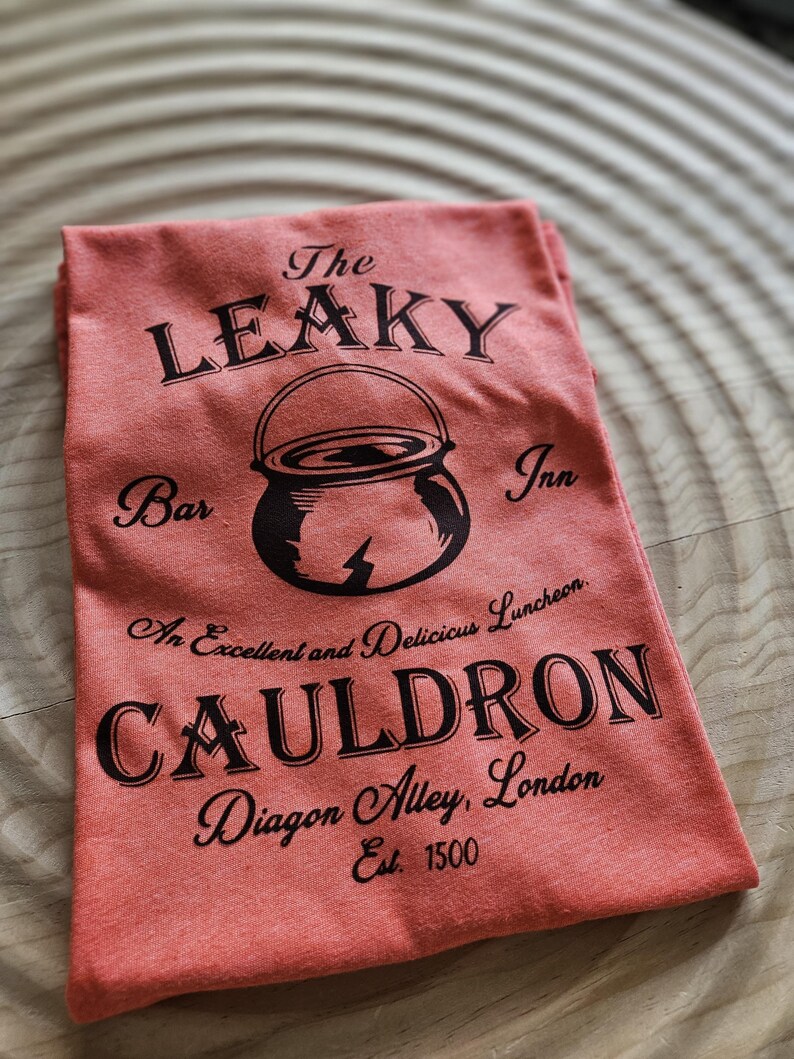 May include: A red t-shirt with a black graphic of the Leaky Cauldron, a bar and inn in Diagon Alley, London. The text reads "The Leaky Cauldron, Bar Inn, An Excellent and Delicious Luncheon, Cauldron, Diagon Alley, London, Est. 1500."