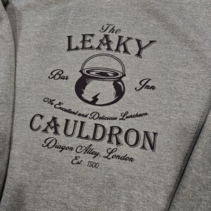 May include: A gray sweatshirt with a black graphic print of the Leaky Cauldron, a fictional pub from the Harry Potter series. The graphic includes the text "The Leaky Cauldron", "Bar", "Inn", "An Excellent and Delicious Luncheon", "Cauldron", "Diagon Alley, London", and "Est. 1500".