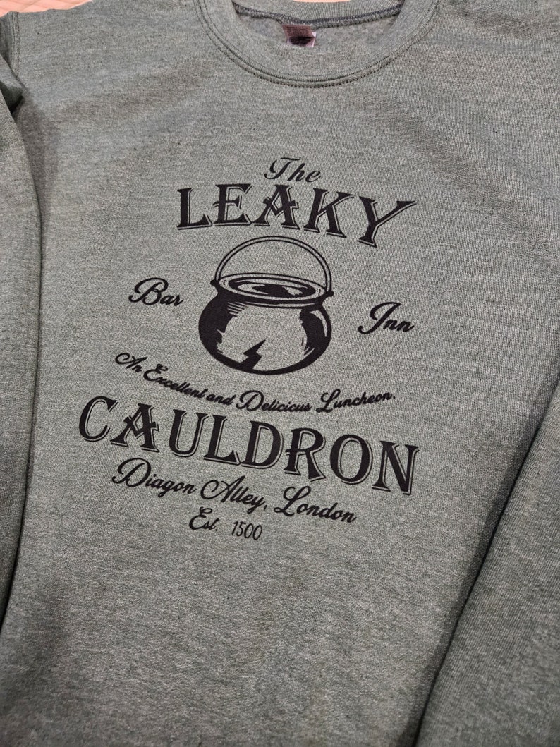 May include: A gray sweatshirt with a black graphic print of the Leaky Cauldron, a fictional pub from the Harry Potter series. The graphic includes the text "The Leaky Cauldron", "Bar", "Inn", "An Excellent and Delicious Luncheon", "Cauldron", "Diagon Alley, London", and "Est. 1500".