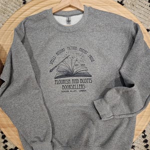 May include: A heather gray sweatshirt with a crew neck and long sleeves. The front features a black graphic of an open book with a wand and the text "SPELLS HISTORY FICTION POETRY PROSE" and "FLOURISH AND BLOTTS BOOKSELLERS DIAGON ALLEY, LONDON."