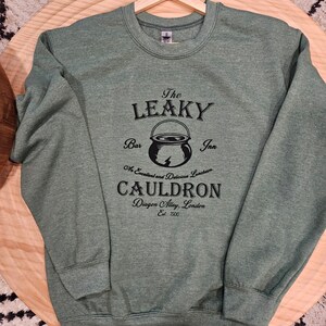 May include: Green sweatshirt with a black graphic of The Leaky Cauldron, a bar and inn in Diagon Alley, London. The graphic includes a cauldron with a lightning bolt and the text "An Excellent and Delicious Lunchroom".