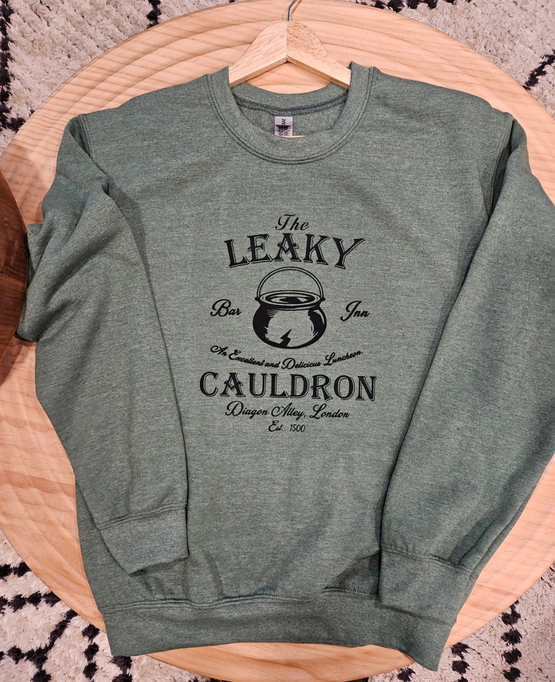 May include: Green sweatshirt with a black graphic of The Leaky Cauldron, a bar and inn in Diagon Alley, London. The graphic includes a cauldron with a lightning bolt and the text "An Excellent and Delicious Lunchroom".