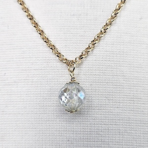 May include: A gold-colored chain necklace with a faceted, clear crystal pendant. The pendant is a round, multi-faceted bead that reflects light. The necklace is displayed against a white background.