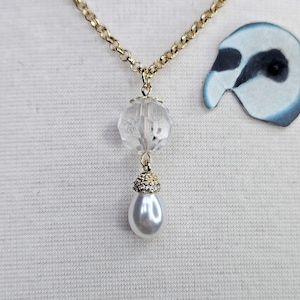 Puede incluir: Un collar dorado con un colgante de bola de cristal transparente y facetado y una gota de perla de imitación en forma de pera. El collar se muestra sobre un fondo de tela blanca.