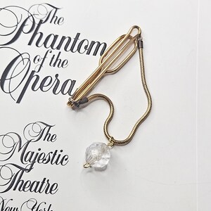 May include: A gold-toned tie clip with a chain and a clear, faceted crystal pendant. The clip is on a white surface with the words "The Phantom of the Opera" and "The Majestic Theatre New York" in black script.