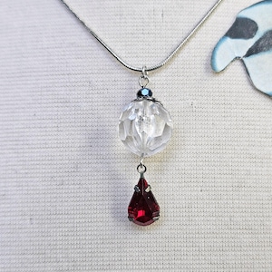 Puede incluir: Un collar plateado con un colgante de orbe claro y facetado, con una pequeña cuenta azul. Debajo del orbe cuelga un colgante en forma de lágrima, de color rojo intenso. El collar se muestra sobre un fondo blanco.