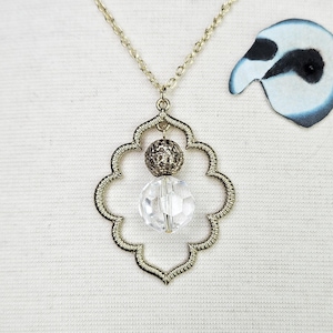 Puede incluir: Un collar dorado con un colgante decorativo con un diseño de filigrana. El colgante contiene una cuenta de cristal transparente facetada y una cuenta gris oscuro texturizada. El collar tiene una cadena delicada.