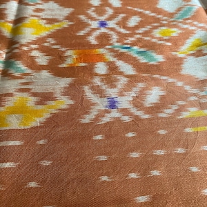以下が含まれることがあります： 白と黄色のイカット模様の入った茶色の布。布は上品な光沢と柔らかな質感があります。