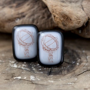 Pode incluir: Dois botões de punho quadrados de vidro com bordas pretas. Cada botão de punho apresenta um fundo branco com um desenho de linha marrom de uma figura segurando um globo.