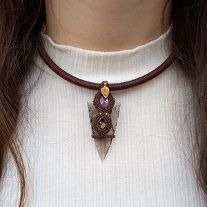 May include: A brown leather cord choker necklace with a pendant featuring a clear, grey, and purple arrowhead with gold accents.