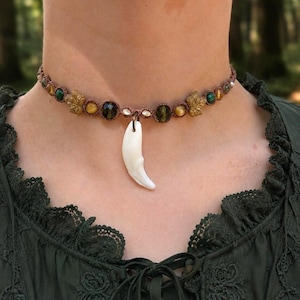 May include: A brown macrame choker necklace featuring a central white curved tooth-shaped pendant. The band is decorated with alternating round green and golden-brown beads, interspersed with small tan leaf-shaped charms and tiny white accents.