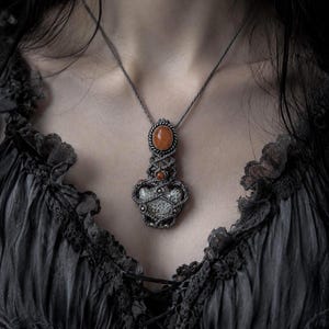Può includere: Una collana con ciondolo in macramè caratterizzata da un cordino grigio scuro, una pietra ovale arancione liscia in alto, una perlina arancione più piccola sotto e una pietra testurizzata dal motivo chiaro alla base, racchiusa in nodi grigi.