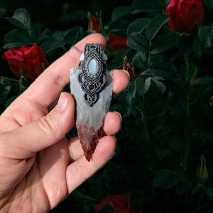 Puede incluir: Un colgante de punta de flecha gris y rojo con un diseño de macramé y una piedra blanca en el centro. El colgante se sostiene en una mano sobre un fondo de hojas verdes y rosas rojas.