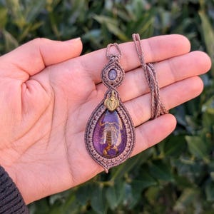 Op de afbeelding: Een handgemaakte macramé ketting met een druppelvormige hanger. De hanger heeft een paars hars midden met een bewaard gebleven insect. De ketting heeft een bruin koord en ingewikkelde macramé details in bruin- en paars tinten. Een uniek, door de natuur geïnspireerd sieraad.