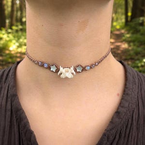 May include: A brown macrame choker necklace featuring a central white animal vertebra bone, flanked by two light-colored star-shaped beads and smaller round iridescent moonstone-like beads integrated into the woven cord band.