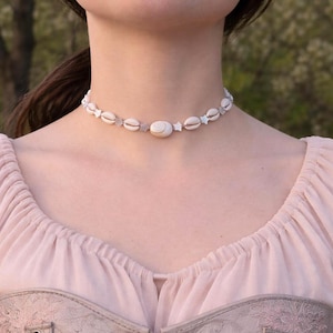 Puede incluir: Un delicado collar gargantilla con una gran concha ovalada central, rodeada de conchas más pequeñas y cuentas en forma de estrella. El collar está ensartado en un cordón fino y claro, creando una estética playera y bohemia. El collar lo lleva una persona.