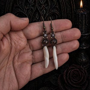 Könnte beinhalten: Handgefertigte Ohrringe mit Makramee-Design, braunen Akzenten, kleinen Edelsteinen und langen, weißen, zahnförmigen Anhängern. Die Ohrringe haben eine gotische oder böhmische Ästhetik.