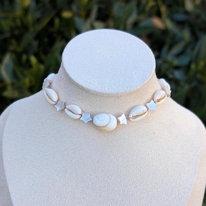 Puede incluir: Un collar de conchas blancas, con una concha grande central y detalles en forma de estrella. El collar se exhibe sobre un busto de maniquí blanco. El fondo es verde borroso, evocando un tema costero o marino.