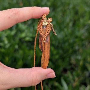 Può includere: Collana con pendente fatto a mano con base in legno, dettagli in macramè e una gemma centrale. Il pendente presenta dettagli in metallo color oro e un cordino abbinato. I materiali naturali creano un'estetica unica e terrosa.