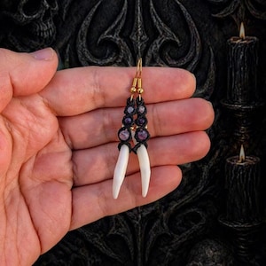 May include: A pair of macrame earrings featuring white, elongated pendants, accented with purple beads and black macrame knots. The earrings have gold-colored hooks. The background is dark with a skull design and lit candles.