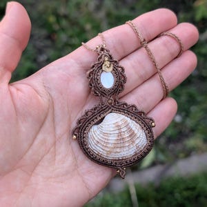 Könnte beinhalten: Eine braune Makramee-Kette mit einem Muschelanhänger. Die Muschel ist weiß mit braunen Streifen und von braunem Makramee umgeben. Die Kette ist an einer braunen Schnur.