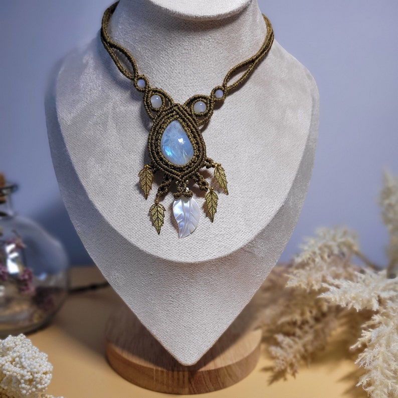 khaki bzw. olivgrüne Makramee Kette mit einem blau schimmernden Regenbogen Mondstein, weißen Achat Perlen, einem geschnitzten Perlmutt Blatt, und kleinen goldenen Blatt Anhängern, an einer beigen Schmuckpuppe