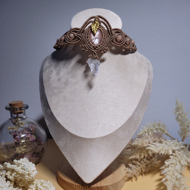 Makramee Kette in der Farbe altrosa mit einem Pfirsich Mondstein, einer klaren Bergkristall Pfeilspitze, einem Messing Blatt Anhänger und Pfirsich Mondstein Perlen, an einer beigen Schmuckpuppe.