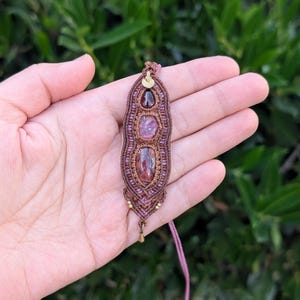 May include: A handcrafted macrame pendant necklace featuring three gemstones in shades of red and pink. The pendant is intricately woven with brown and purple threads, accented with gold-colored beads and a delicate chain.