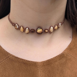 Può includere: Una collana girocollo in macramè marrone con una serie di pietre naturali. Le pietre sono nelle tonalità del beige, marrone e giallo. La collana è indossata intorno al collo, mettendo in risalto l'intricato lavoro di macramè e i toni della terra delle pietre.