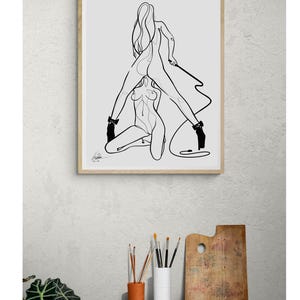 May include: A framed black and white line art print depicting two figures in a sensual pose. The artwork features minimalist lines, high heels, and a whip. The print is displayed above a wooden table with art supplies.