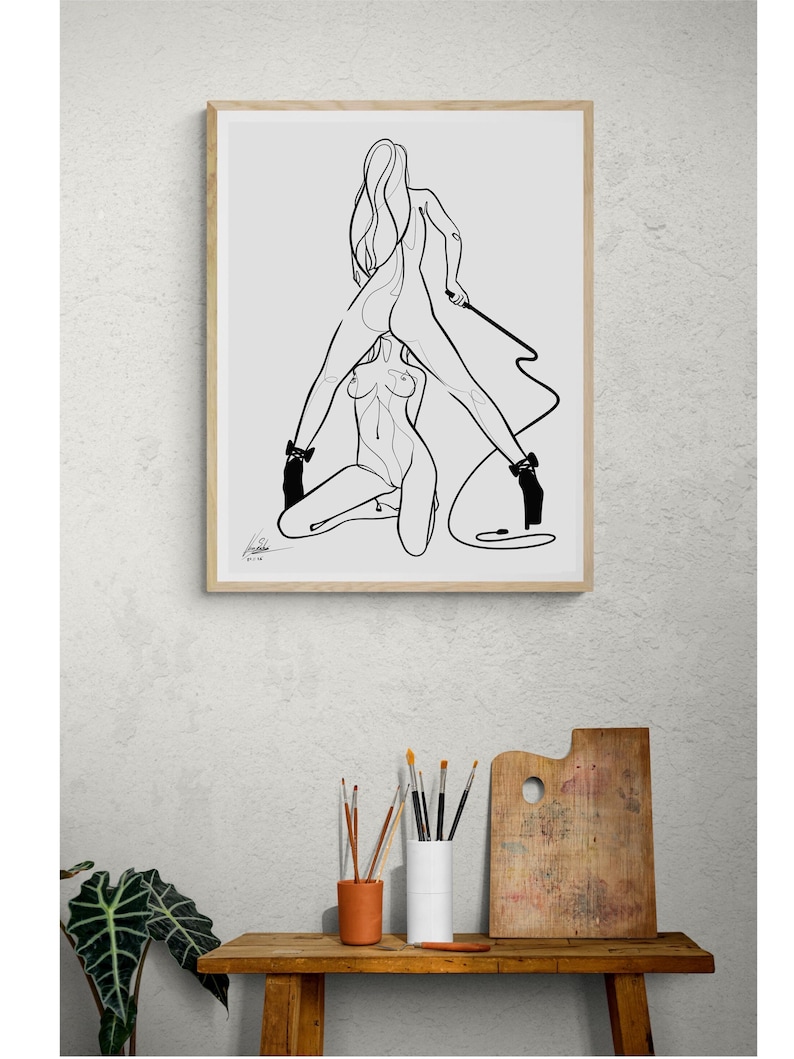 May include: A framed black and white line art print depicting two figures in a sensual pose. The artwork features minimalist lines, high heels, and a whip. The print is displayed above a wooden table with art supplies.