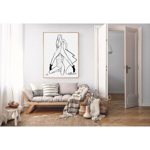 May include: A framed black and white line art print depicting two figures in an embrace. The artwork hangs above a wooden sofa with pillows and a striped blanket. The room has a neutral color palette with natural textures.