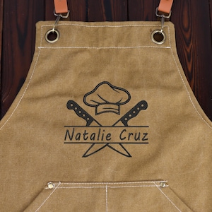 May include: Tan canvas apron with brown leather straps and metal hardware. The apron features a chef hat and crossed knives graphic with the name "Natalie Cruz" in black. A pocket is visible at the bottom.